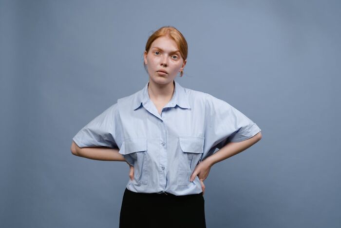 Young woman with red hair looking annoyed with hands on hips against a plain blue background, showing Karen attitude.