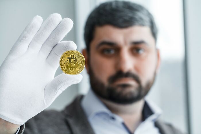 Man wearing white glove holding a Bitcoin coin close to the camera, illustrating people falling for scams and consequences.