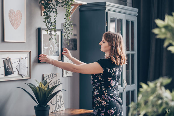 Woman adjusting framed photos on wall in a home, illustrating jealous wife reacting to hubby&rsquo;s mom having pics of his ex.
