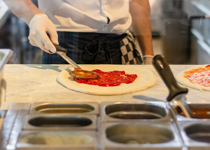 Restaurant owner preparing pizza dough with tomato sauce in kitchen amid controversy for insulting tourists sharing pizzas. Restaurant owner preparing pizza dough with tomato sauce in kitchen amid controversy for insulting tourists sharing pizzas.