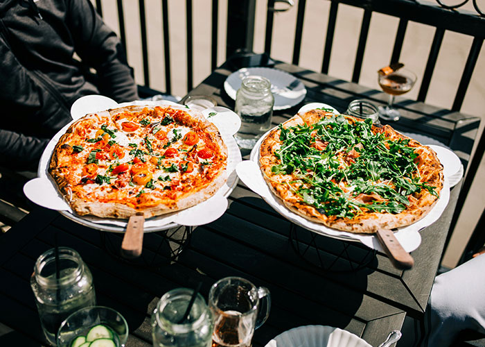 Two large pizzas with fresh toppings on wooden paddles at an outdoor table in a restaurant setting. Two large pizzas with fresh toppings on wooden paddles at an outdoor table in a restaurant setting.