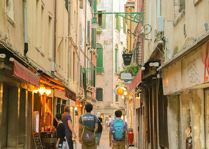 Tourists walking down a narrow European street near restaurants causing restaurant owner backlash over pizza sharing. Tourists walking down a narrow European street near restaurants causing restaurant owner backlash over pizza sharing.