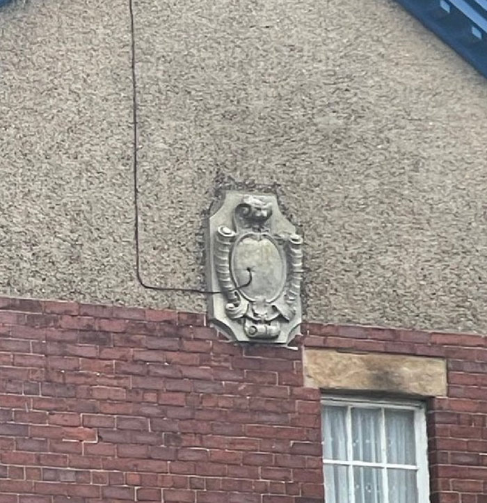 Uneven brick and stucco wall with a decorative but oddly placed architectural plaque near a window, showcasing design and architecture flaws.