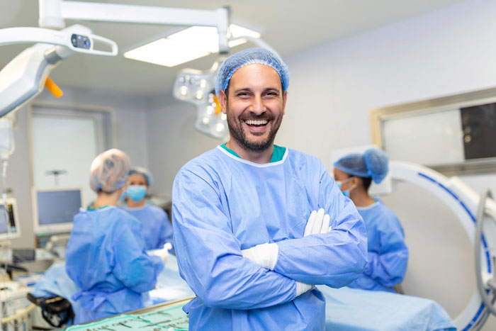 Smiling male surgeon in scrubs and cap stands confidently in operating room during cosmetic surgery mole removal procedure.