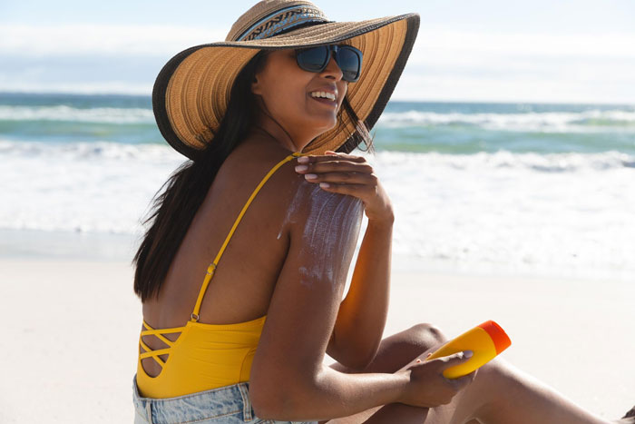 Young woman at the beach applying sunscreen, smiling and wearing a sunhat and sunglasses on a sunny day.