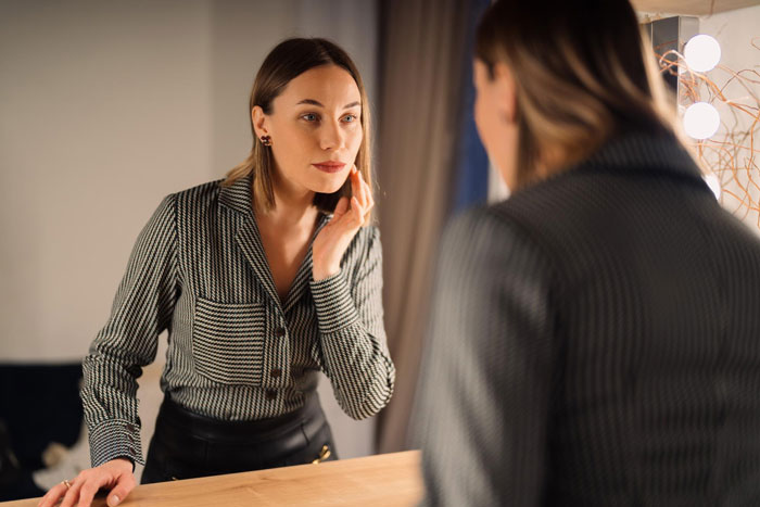 Young woman looking at her face in the mirror, reflecting on plastic surgery and appearance concerns in a dimly lit room.