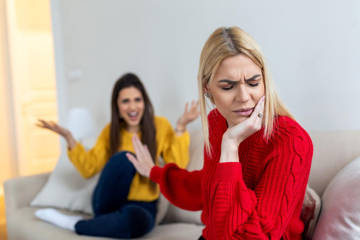 Two sisters in a living room, one looks upset refusing a plastic surgery gift, the other gesturing angrily.