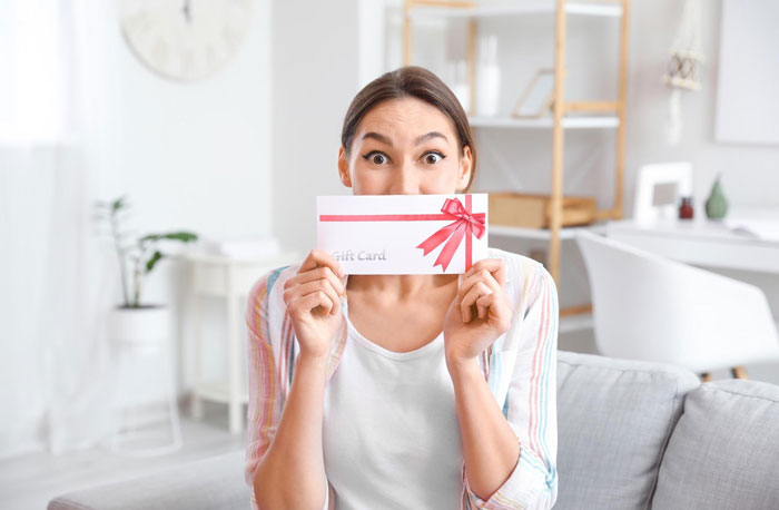 Young woman holding a plastic-surgery gift card with a surprised expression in a bright living room setting