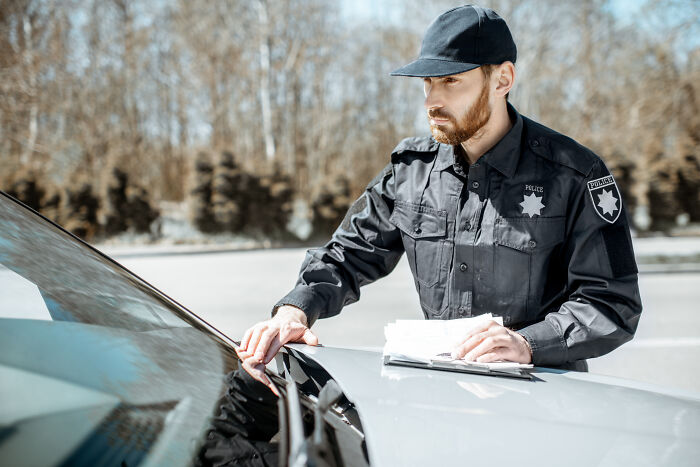 Police officer in black uniform inspecting damage on a car, illustrating dumb drivers instantly getting what they deserved.