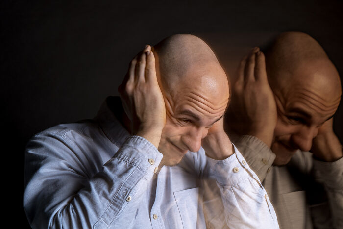 Man with bald head covering ears, appearing distressed, with a shadowed reflection against a dark background, creepy cases concept.