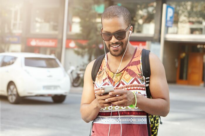 Young man wearing sunglasses and colorful tank top smiling at phone, representing people who fell for scams.