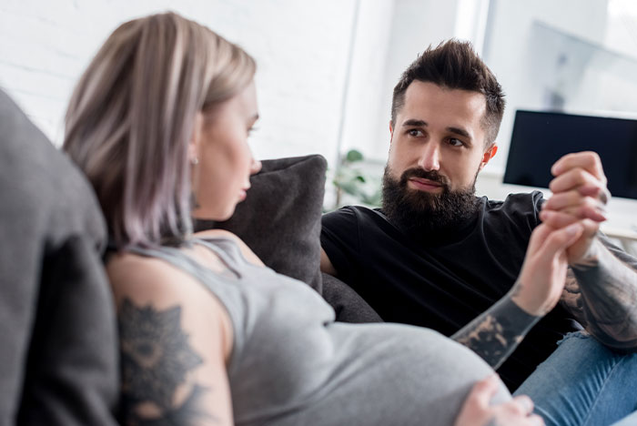 Pregnant girlfriend sitting on couch looking away while boyfriend with beard tries to talk, showing relationship tension.