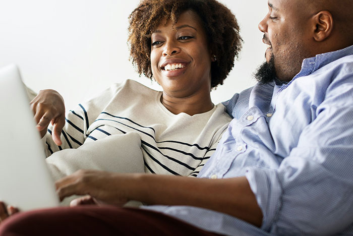 Pregnant girlfriend smiling and sitting on a couch with her partner, using a laptop together in a cozy setting.