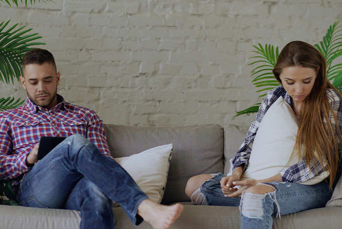 Pregnant girlfriend sitting apart on couch with folded arms, refusing to move together, while boyfriend looks at tablet.