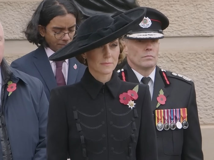 Princess Kate attending a formal event wearing a black hat and coat, with officials in uniform nearby.