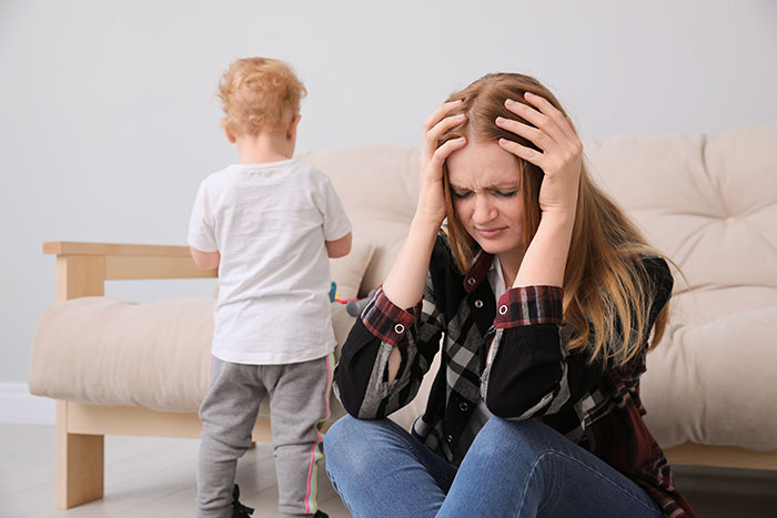 Stressed mother holding her head while child stands nearby, illustrating parents and children share reasons parents were unhappy.