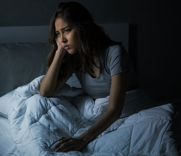 Woman awake in bed at night, troubled by roommate&rsquo;s snoring, unable to sleep under dim bedroom lighting.