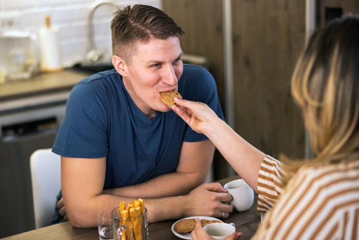Woman feeding boyfriend a cookie at kitchen table, showcasing perfect secret cookies and a joyful moment together. Woman feeding boyfriend a cookie at kitchen table, showcasing perfect secret cookies and a joyful moment together.