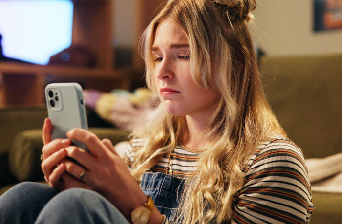 Teen sitter looking concerned at phone while seated indoors, reflecting on caring for a nonverbal bedridden boy.
