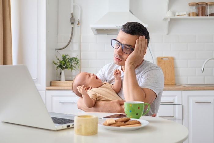 Frustrated man holding baby while working on laptop at home, illustrating babysitting on vacation dilemma.