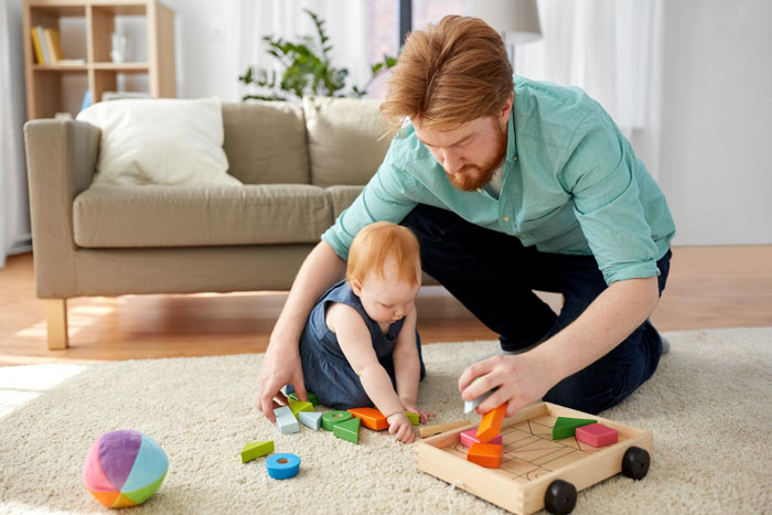 Man spending time with toddler playing with wooden toys indoors, illustrating babysitting on vacation concerns.