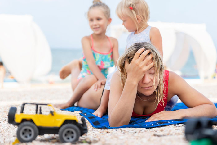 Frustrated woman overwhelmed by kids on beach, illustrating conflict with babysitting on vacation and personal time.