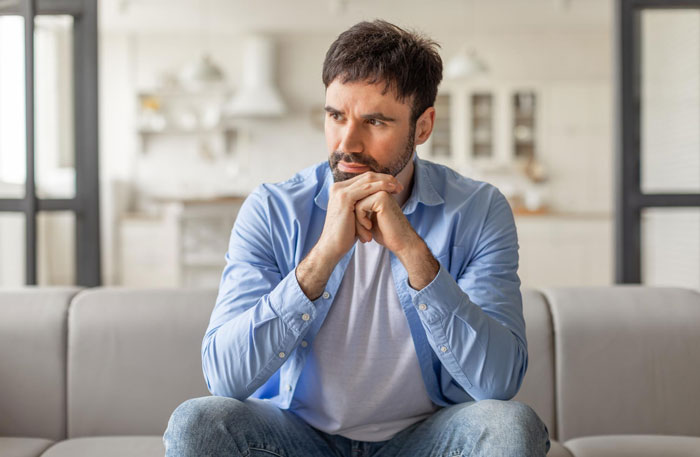 Man in a blue shirt sitting on a couch looking thoughtful and concerned, reflecting on cosmetic surgery recovery help refusal.