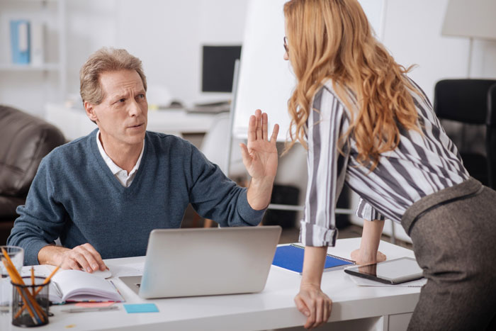 Man refusing to train replacement without pay, raising hand in disagreement during office conversation with woman.