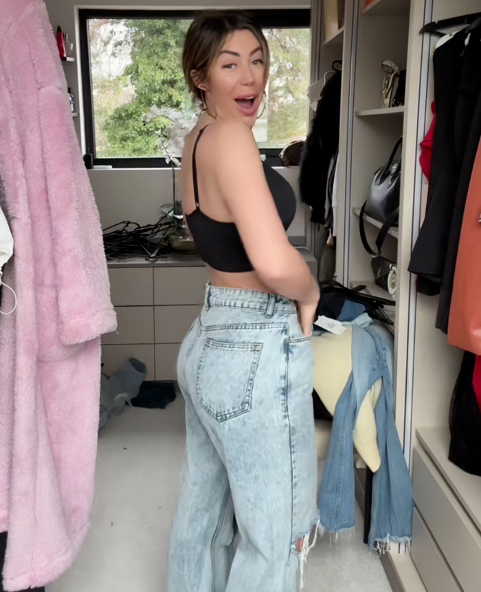 Woman smiling in a black top and jeans in a closet, related to expert surgeon revealing regretted plastic surgery.