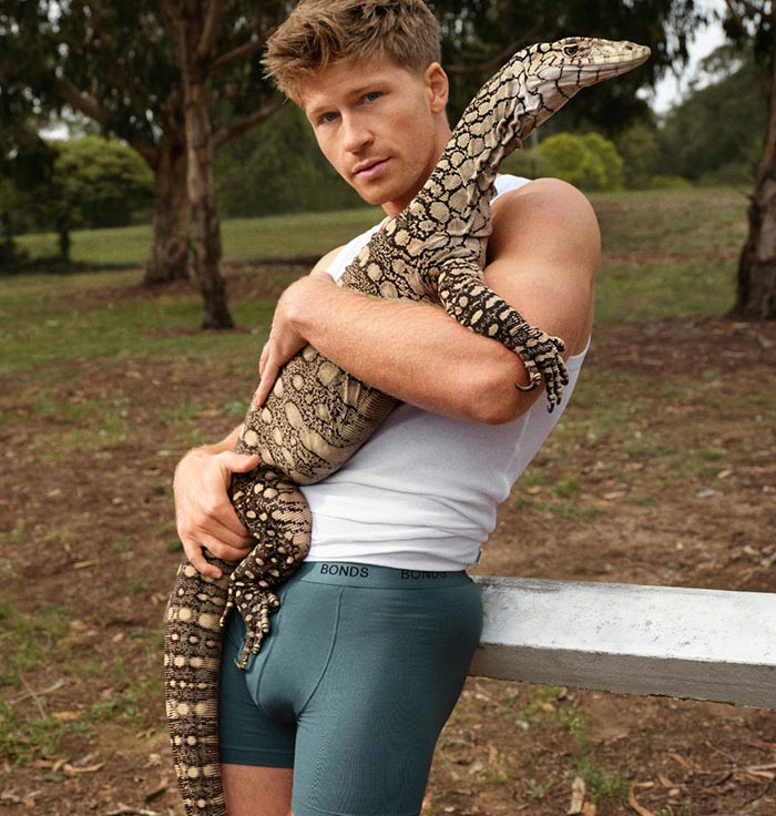 Shirtless man wearing Bonds underwear with a large snake draped over his shoulders in an outdoor rural setting.