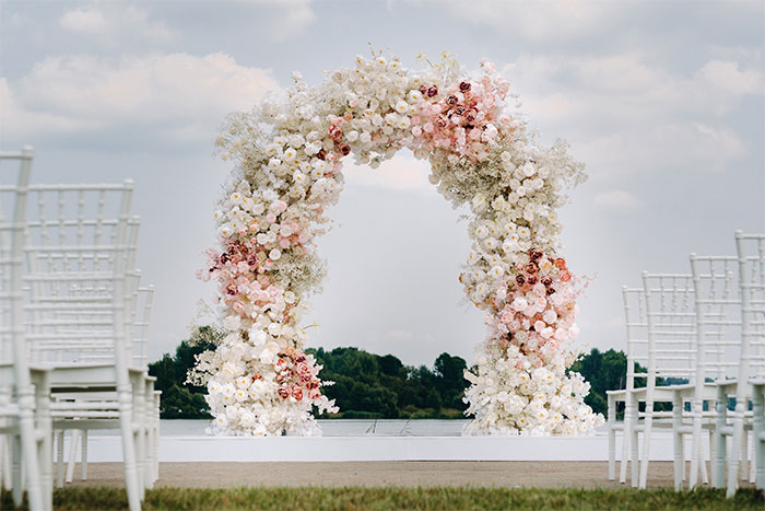 Floral wedding arch setup by a lake with rows of white chairs for a marriage proposal event outdoors. Floral wedding arch setup by a lake with rows of white chairs for a marriage proposal event outdoors.