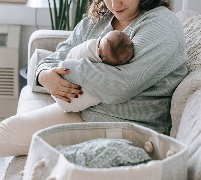 Mother sitting on couch holding newborn baby wrapped in white blanket near a bassinet, family moment after 30 years and 4 kids. Mother sitting on couch holding newborn baby wrapped in white blanket near a bassinet, family moment after 30 years and 4 kids.