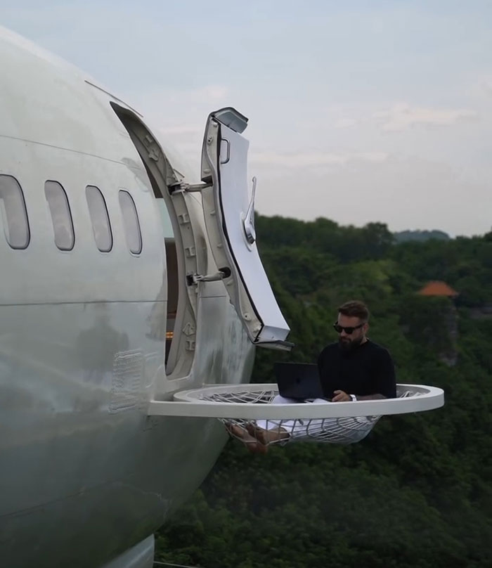 Man working on laptop sitting in hanging net platform outside airplane door floating above forest, crypto scammer concept.