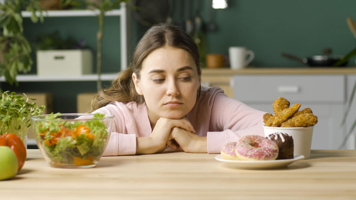 Young woman in kitchen choosing between fast food and healthy salad, reflecting on strict diet and eating disorder triggers.