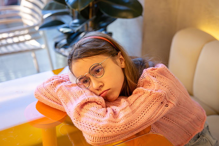 Sad young girl in pink sweater resting head on folded arms, feeling ignored and hurt due to family work priorities.