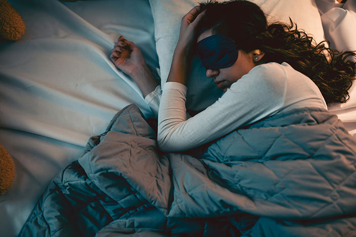 Woman wearing a sleep mask lying in bed under a blanket, depicting the scariest things people have woken up to.