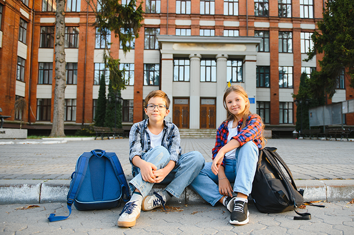 Two stepchildren sitting outside a boarding school with backpacks, waiting for a woman&rsquo;s request.