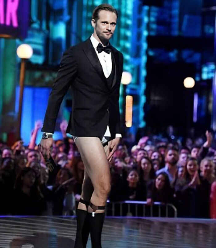 Alexander Skarsg&aring;rd in a tuxedo jacket and bow tie with unique legwear on stage in front of a large audience at an event.