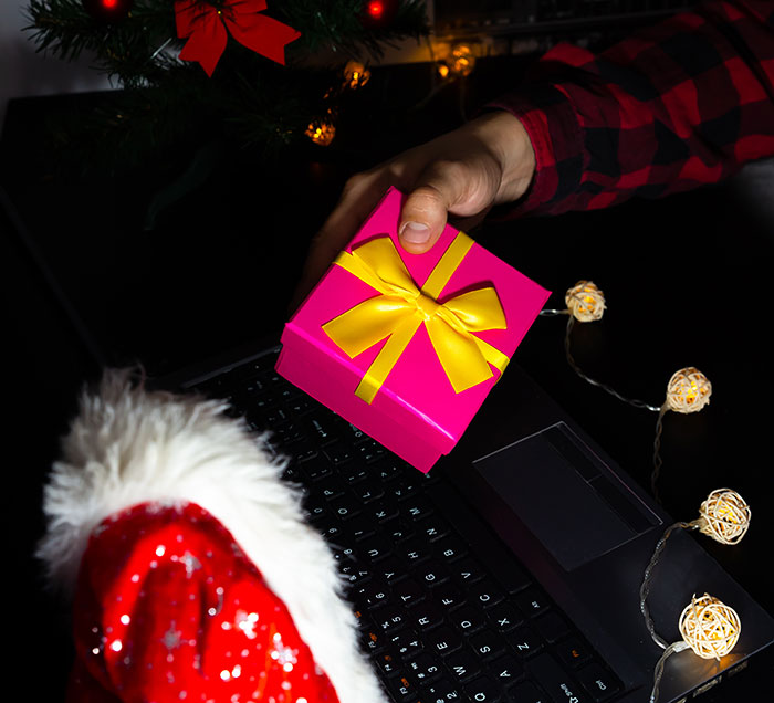 Hand holding pink gift box with yellow ribbon near laptop and Christmas decorations for Secret Santa organizer event.