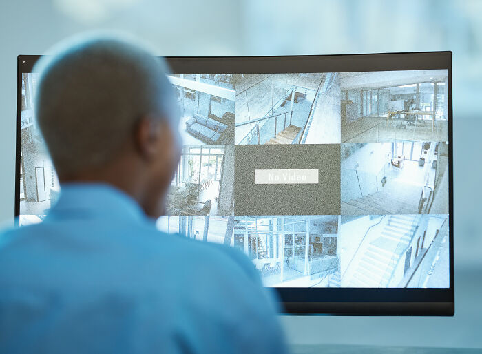 Man monitoring multiple surveillance cameras on a curved screen, capturing various empty indoor scenes and staircases.