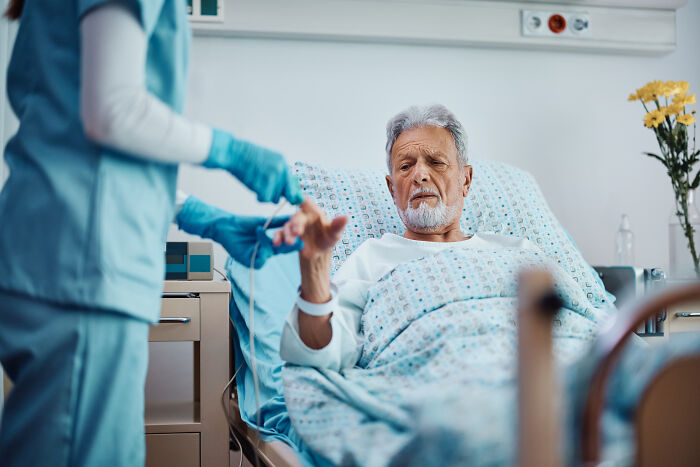 Elderly patient in hospital bed with nurse checking his hand, illustrating eerie patient stories that haunt doctors.