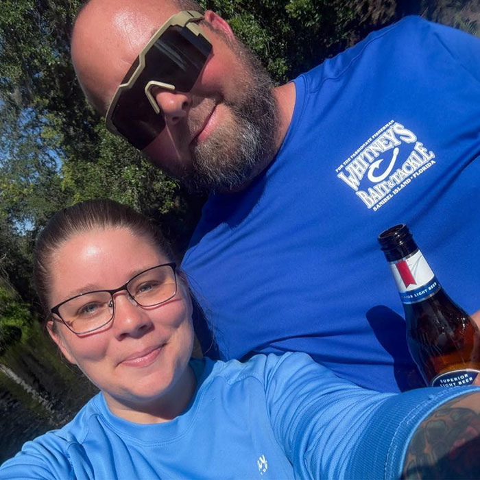 Couple outdoors wearing blue shirts and sunglasses, enjoying a sunny day near trees with a beer bottle visible.