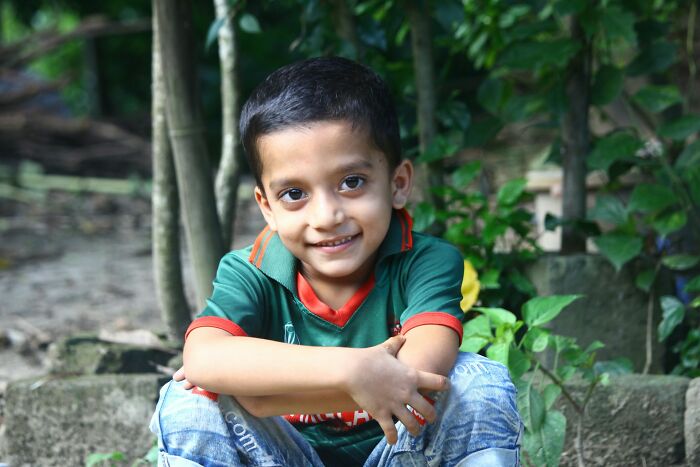 Young boy smiling outdoors sitting on stone steps with greenery in background, capturing moments of funny and accidental hurt.