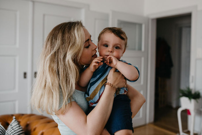 Woman feeling sick at home holding baby, appearing to ask husband for money in a cozy living room setting