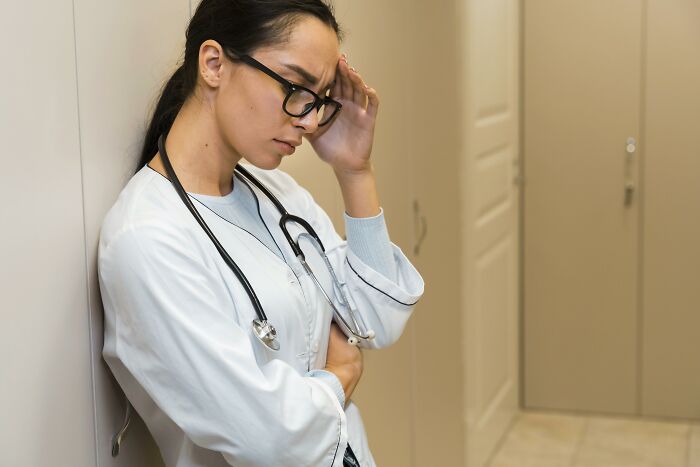 Stressed female doctor wearing glasses and a stethoscope leaning against wall reflecting on creepy medical cases.