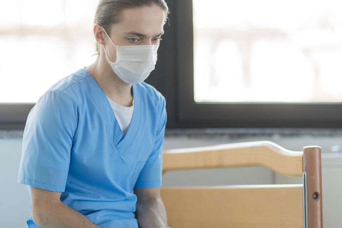 Young nurse wearing a mask and blue scrubs, sitting thoughtfully by a hospital window during a quiet moment.