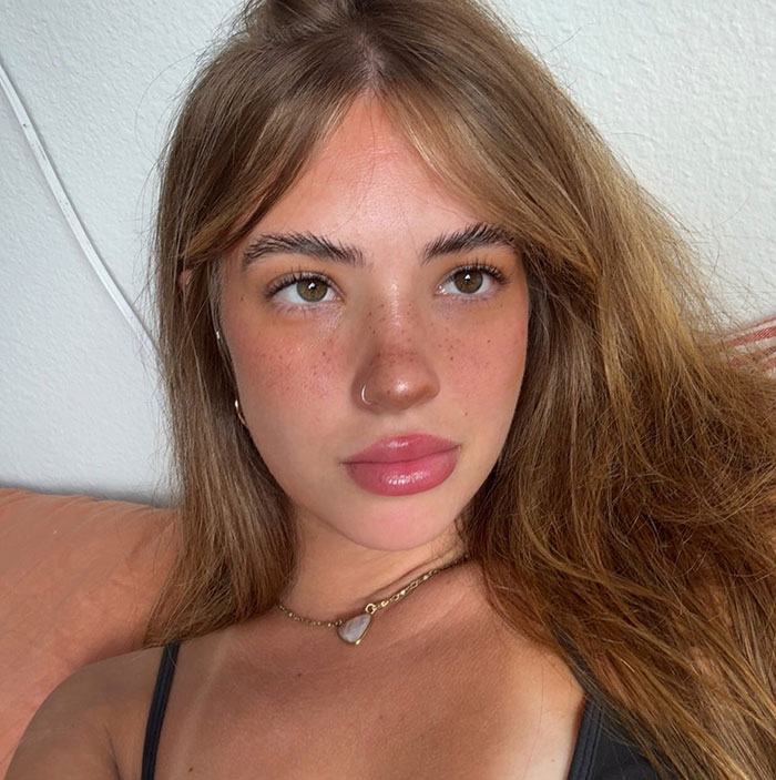 Young woman with tattooed freckles and long hair, looking thoughtfully to the side in natural lighting indoors.