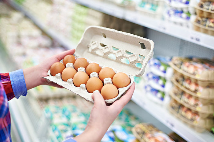 Person holding a carton of brown eggs in a grocery store aisle representing the college experience in America