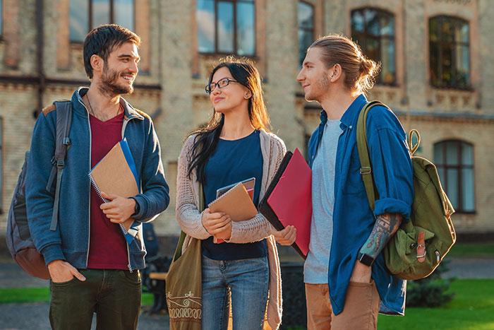 Three college students with backpacks and notebooks smiling outside a historic campus building, capturing the college experience.