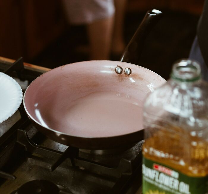 Close-up of a pink frying pan on a gas stove next to a bottle of sunflower oil in a home kitchen setting.